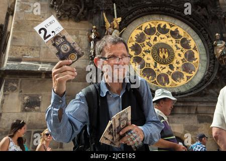 Der ältere Straßenhändler verkauft Postkarten vor der berühmten astronomischen Uhr (orloj) auf dem Turm des Alten Rathauses (Staroměstská radnice) auf dem Altstädter Ring (Staroměstské náměstí) in Prag, Tschechien. Stockfoto