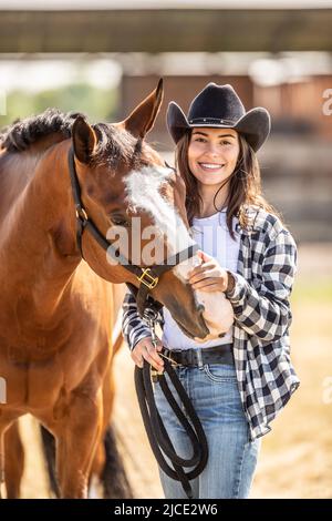 Eine gut aussehende junge Frau mit Cowboyhut streichelt ein Pferd auf Koteletts, während sie die Kamera anlächelt. Stockfoto