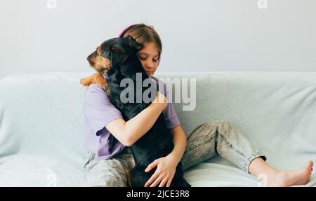 Fröhliches Teenager-Mädchen in lila T-Shirt sitzt auf dem Sofa, hört Musik hat Spaß mit dem Hund spielen Stockfoto