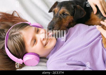 Fröhliches Teenager-Mädchen im lila T-Shirt in rosa Kopfhörern hört Musik und hat Spaß beim Spielen mit Hund Stockfoto