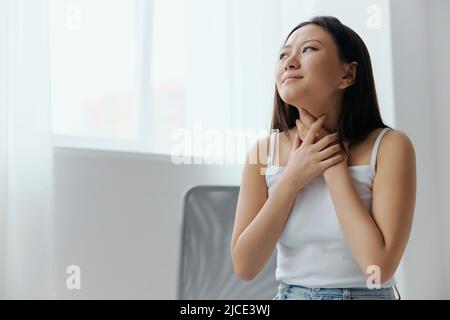 Halsschmerzen vergrößerte Lymphknoten. Trauriges Leiden gebräunt schöne junge asiatische Frau berühren Hals zu Hause innen Wohnzimmer. Schlechte Verletzungen Stockfoto