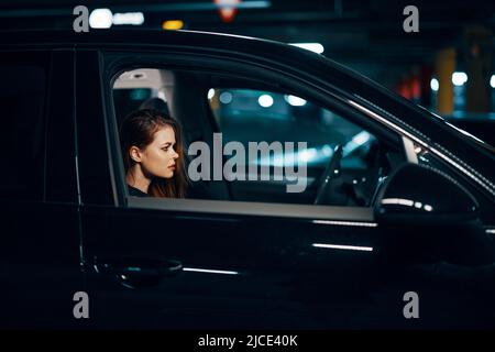 Horizontales Foto von der Seite, nachts, von einer Frau, die in einem schwarzen Auto sitzt und nachdenklich nach vorne schaut Stockfoto