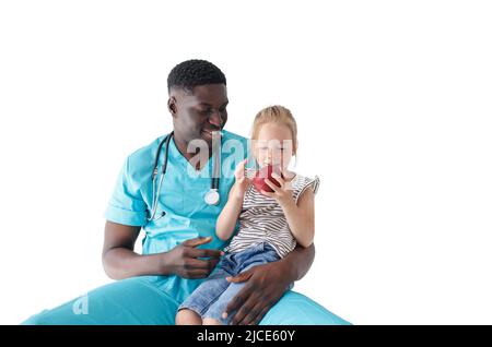 Ein afroamerikanischer Kinderarzt behandelt einem kleinen Mädchen, das auf dem Schoß des Arztes auf einem weißen, isolierten Hintergrund sitzt, einen Apfel Stockfoto
