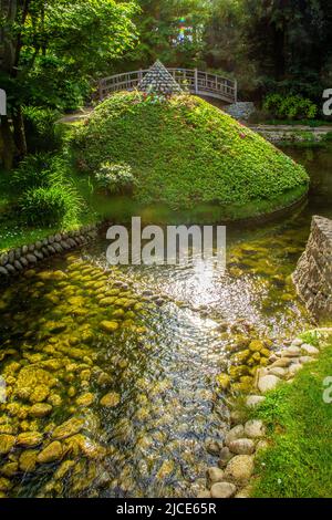 Der Teich mit Steinbänken und einer Art Azaleen Büschen Pyramide und japanische Brücke dahinter und Sonne glänzte in Wasserspiegelungen in Albert Kahn Stockfoto