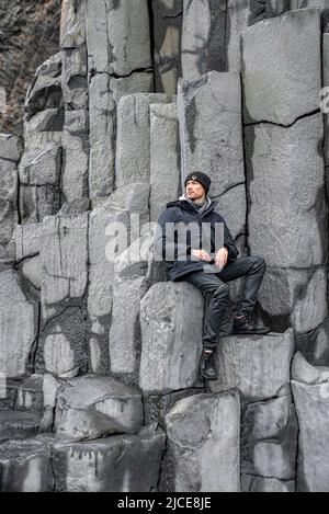 Nachdenklicher Tourist, der auf Basaltsäulen am Strand von Reynisfjara sitzt Stockfoto