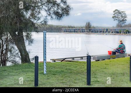 Ein Mann fischt von einem Kai in der Nähe einer Flutmarkierung und Ferienhütten, die am Ufer des Lake Conjola an der australischen Südküste, NSW, gebaut wurden Stockfoto