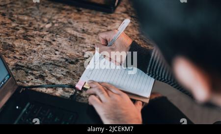 Person, die auf einem Notizbuch auf der Theke seines Schreibtisches schreibt Stockfoto