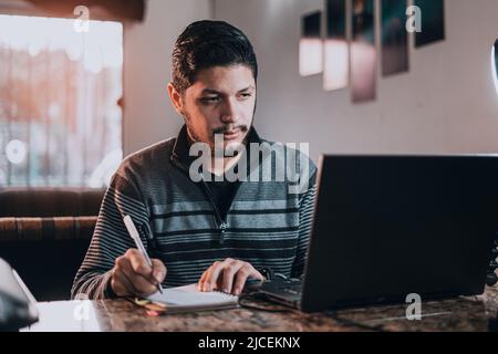 Junger Mann, der online auf einem Laptop studiert, sitzt hinter seinem Schreibtisch im Wohnzimmer Stockfoto