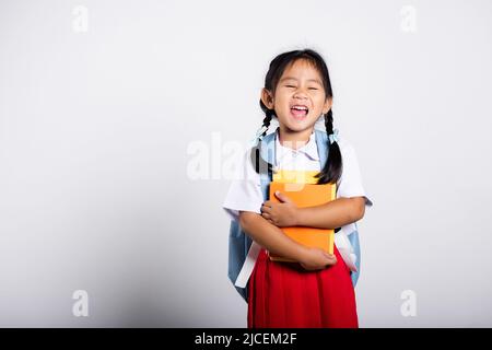 Asiatische liebenswert Kleinkind lächelnd glücklich tragen Student thai uniform roten Rock stehen halten oder umarmende Buch im Studio geschossen isoliert auf weißem Hintergrund, Portr Stockfoto