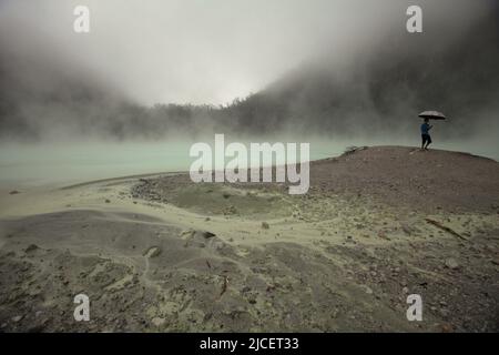 Ein Besucher, der einen Regenschirm trägt, während er auf sandiger Landschaft im Hintergrund des nebligen Krater des Mount Patuha spaziert, der im Volksmund als Kawah Putih (weißer Krater) in Ciwidey, Bandung, West Java, Indonesien, bekannt ist. Stockfoto