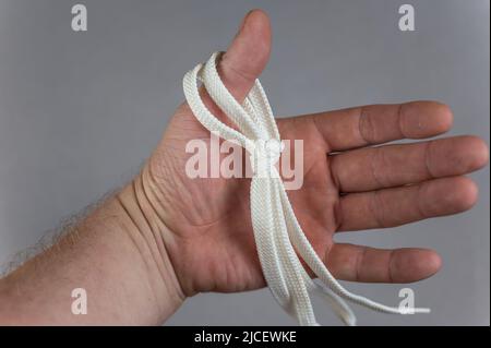 Ein Mann, der eine weiße Trageschlaufe in der Hand hält. Grauer Hintergrund. Flacher Schnürsenkel, der in offener Handfläche am Daumen hängt. Reifer Mann. Nahaufnahme. Selektiver Fokus. Stockfoto