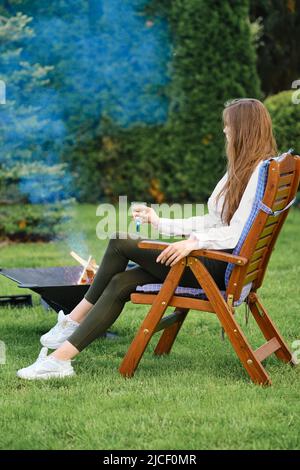 Junge Frau, die in einem Liegestuhl auf dem Hinterhof neben einem Grill ruht, Blick auf das Profil Stockfoto