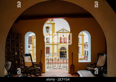 Blick von einem Zimmer mit offener Tür und Fenstern auf eine Kolonialkirche in Santa Cruz de Mompox, Kolumbien, Weltkulturerbe Stockfoto