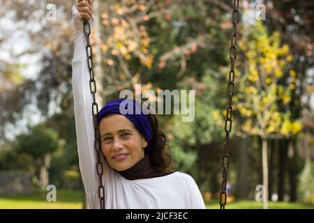 Lächelnde Dame mittleren Alters mit purpurfarbenem Schal auf dem Kopf hält die Schaukelkette im Park und grüne Bäume im Hintergrund. Ruhige reife Frau auf dem Spielplatz Stockfoto