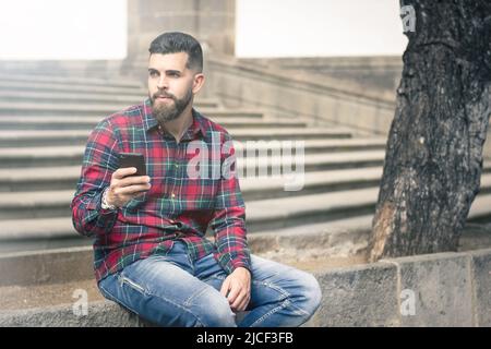 Junger Hipster mit Zellular, der in der Altstadt an einer Steinmauer an einem Baum sitzt. Nachdenklicher, gutaussehender Mann, der Smartphone auf der Straße hält. Lichteffekt Stockfoto