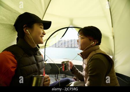 Zwei fröhliche junge asiatische Frauen sitzen im Zelt zusammen, hören Musik in den Kopfhörern und trinken Tee oder Kaffee Stockfoto