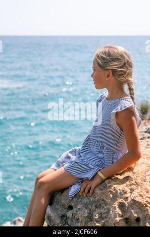 Kleine Kinder Mädchen Reisende acht Jahre alt sitzt auf einem Felsen oder einem Stein an der Küste des Meeres und bewundert die inspirierende Aussicht auf das Wasser und Stockfoto