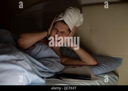 Junge Frau mit einem weißen Handtuch, das um den Kopf gewickelt ist und auf dem Bett liegt und Kopfschmerzen hat, Ohrenschmerzen, die die Ohren mit den Händen bedecken. Stockfoto