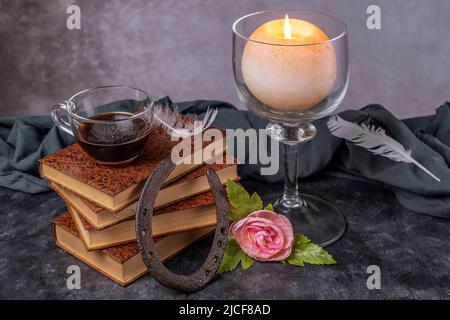 Ein Stapel alter Bücher mit einer Glasschale mit Kaffee darauf, einem Hufeisen, einer Rose und einer brennenden Kerze Stockfoto