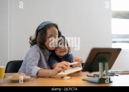 Liebevolle ältere Großmutter umarmt Schultern von kleinen Mädchen Enkelkind helfen bei der Vorbereitung Hausaufgaben Unterstützung geben Ratschläge. Freundliche, ältere Lehrerin Stockfoto