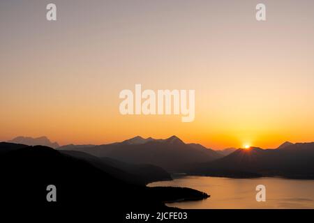 Wolkenloser Sonnenuntergang mit orangefarbenem Abendhimmel über dem Walchensee, im Hintergrund der Simetsberg, das Estergebirge und ganz links die Zugspitze. Stockfoto