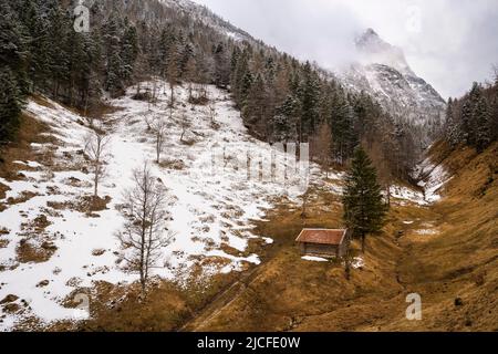 Ein einziger Heuhaufen mit rotem Dach über dem Ferchensee bei Mittenwald in den bayerischen Alpen im Frühjahr. Eine noch teilweise schneebedeckte Bergwiese, mit Wald und Schnee, im Hintergrund die massiven Wettersteinberge mit dem Wetterstein in Wolken und bewölktem Himmel. Stockfoto