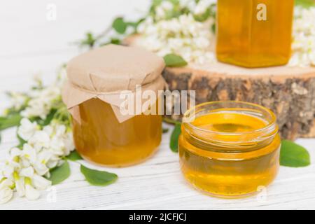 Süßes Honigglas umgab die Akazienblüten des Frühlings. Honig fließt aus einem Löffel in ein Glas. Gläser von klarem frischen Akazienhonig auf Holzgrund. Stockfoto