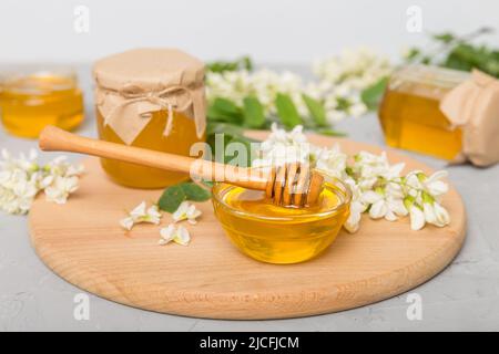 Süßes Honigglas umgab die Akazienblüten des Frühlings. Honig fließt aus einem Löffel in ein Glas. Gläser von klarem frischen Akazienhonig auf Holzgrund. Stockfoto