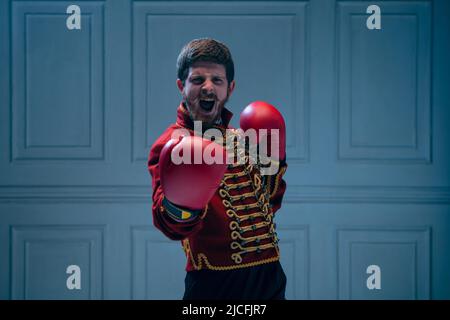 Emotionaler junger Mann wie ein mittelalterlicher Husar, der Boxhandschuhe trägt, isoliert auf dunkelblauem Hintergrund. Retro-Stil, Hobby, Vergleich von Eras Konzept. Stockfoto