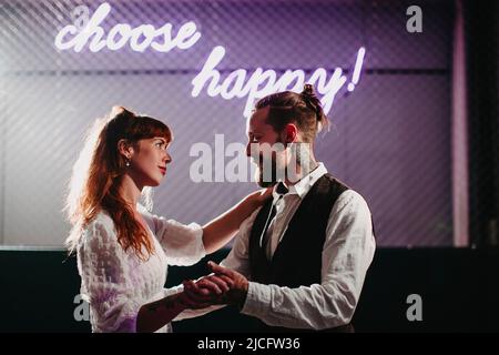 Junges verliebte Paar tanzt unter einem Neonschild „choose happy!“ Stockfoto