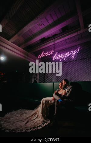 Fröhliche Braut und Bräutigam, Neonschild „choose happy“ Stockfoto