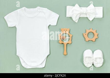 Weißer Baumwoll-Body für Baby-Mockup im Öko-Stil auf mintgrünem Hintergrund. Niedliche Stiefeletten, Stirnband, Holzrassel und Beißringe für Babykleidung moc Stockfoto