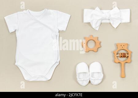 Mockup mit weißem Wickelbody auf weichem beigen Hintergrund. Babyschühchen, Stirnband, Beißringe aus Holz und Rasseln für Babykleidung mockup auf einem Pastell Stockfoto