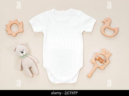 Weißer Babybody für Mockup auf einem pastellfarbenen beigen Hintergrund. Baby-Beißringe aus Holz und gehäkeltes Spielzeug aus Teddybär neben weißem Strampler für Kinder kleiden sich Stockfoto