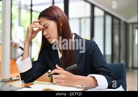 Müde asiatische Geschäftsfrau oder Büroangestellte leidet unter Kopfschmerzen, überfordert, sich aufgeregt und von der Arbeit versagt. Stockfoto