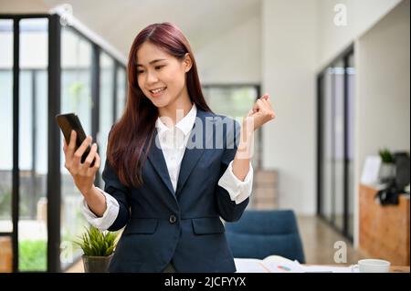 Fröhlich erstaunt erfolgreiche junge asiatische Geschäftsfrau im Büro stehend, mit einem Smartphone, bekommen eine überraschende Nachricht. Stockfoto