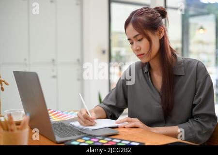 Fokussierte junge asiatische Grafikdesignerin skizziert ihr Design auf einem leeren Notizbuch vor ihrem Laptop an ihrem Schreibtisch. Stockfoto
