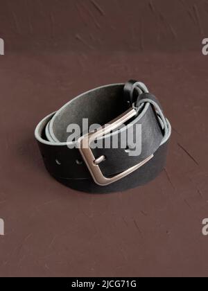 Aufgerollter schwarzer modischer Herren Ledergürtel mit Metallschnalle auf braunem Hintergrund. Männliches Zubehör. Vorderansicht, vertikales Foto. Stockfoto