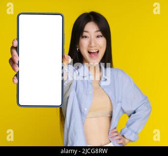 Junge asiatische Mädchen hält Smartphone in der Hand zeigt weißen Bildschirm für Ihre Mock-up-Handy-App-Werbung und aufgeregt Lächeln auf Kamera isoliert auf gelbem Hintergrund. Selektiver Fokus auf das Telefon. Stockfoto