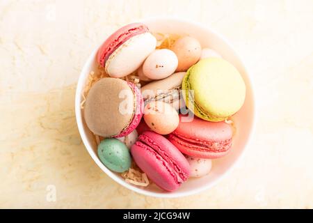 Bunte Makronen und Schokoladeneier in Keramikschale auf beigefarbenem Beton-Hintergrund. Draufsicht, Nahaufnahme, Stillleben. Frühstück, Morgen, Konzept. Stockfoto