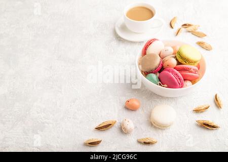 Bunte Makronen und Schokoladeneier in Keramikschale, Tasse Kaffee auf grauem Betongrund. Seitenansicht, Kopierraum, Stillleben. Frühstück, Mo Stockfoto