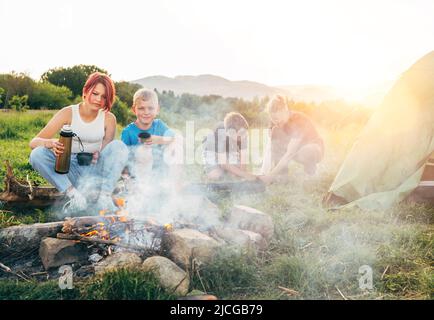 Eine Gruppe lächelnder Kinder in der Nähe eines rauchigen Lagerfeuers, die Tee aus einer Thermoskanne tranken, zwei Brüder bauten das grüne Zelt auf. Glückliche Familie Picknick im Freien Camping aktiv Stockfoto