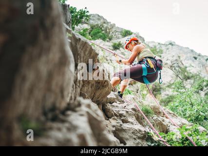 Aktive Klettererin mit Schutzhelm Abseilen von der Felswand mit Seil mit Sicherungsvorrichtung und Klettergurt. Aktive Extremsportzeit Stockfoto