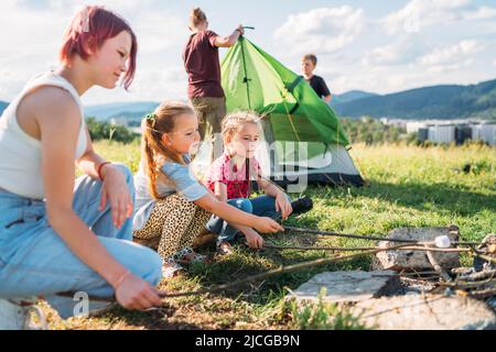 Drei Schwestern lächeln und rösten auf Stöcken über einer Lagerfeuerflamme Sümpfe und Süßigkeiten, während zwei Brüder das grüne Zelt aufschlagen. Glückliche Familie raus Stockfoto