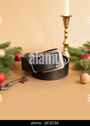 Aufgerollter schwarzer modischer Männer Ledergürtel mit Metallschnalle, Weihnachtstanne, glänzenden Kugeln, goldenem Kerzenständer und Schlüssel auf goldenem Hintergrund. Männlich Stockfoto