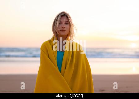 Schöne junge Frau in gelbe Decke gewickelt stehen am Sandstrand bei Sonnenuntergang Blick Kamera. Schöne kaukasische weibliche Person eingewickelt gemütlich karierten Stockfoto