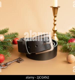 Aufgerollter schwarzer modischer Männer Ledergürtel mit Metallschnalle, Weihnachtstanne, glänzenden Kugeln, goldenem Kerzenständer und Schlüssel auf goldenem Hintergrund. Männlich Stockfoto