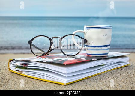 Eine Tasse heißen Tee, Gläser und ein Notizblock mit getrockneten Blättern am Meer. Ruhe am Meer, Entspannung Stockfoto