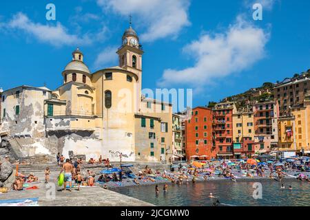 Menschen am Strand als alte Kirche und bunte Häuser unter blauem Himmel auf dem Hintergrund in Camogli, Italien. Stockfoto