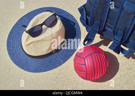 Strohhut, Sonnenbrillen, blauer Rucksack und roter Wasserball auf dem gelben heißen Sand. Konzept für aktiven Urlaub Stockfoto